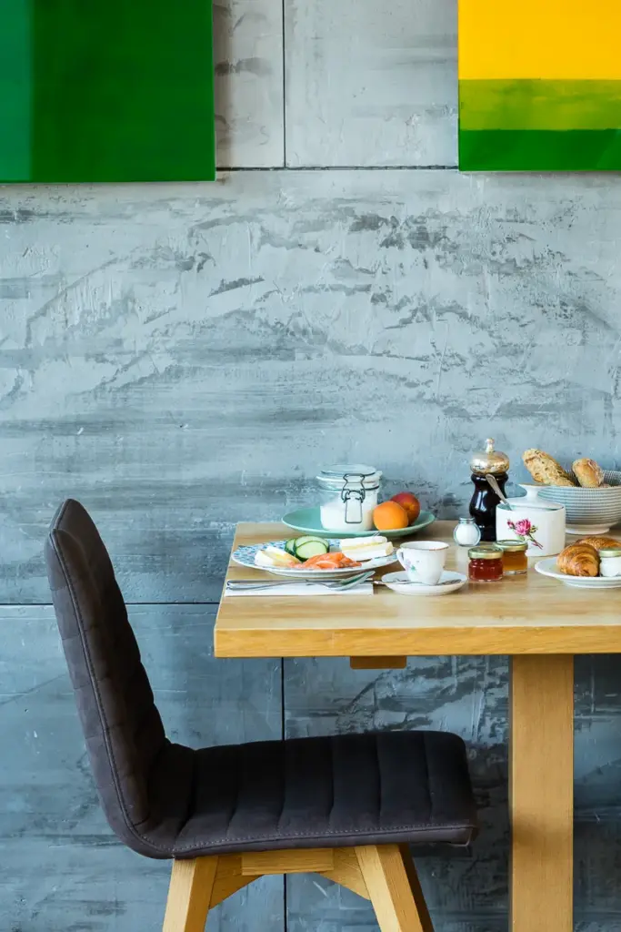 fein gedeckter Fruehstueckstisch vor einer grauen Betonwand mit bunten Bildern